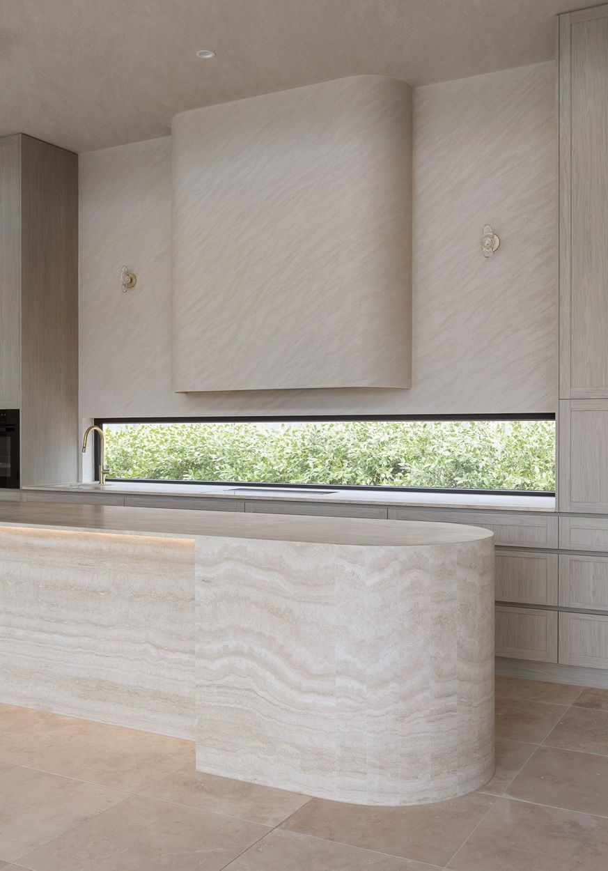 Curved travertine kitchen island in front of a long, low black-framed window above the bench