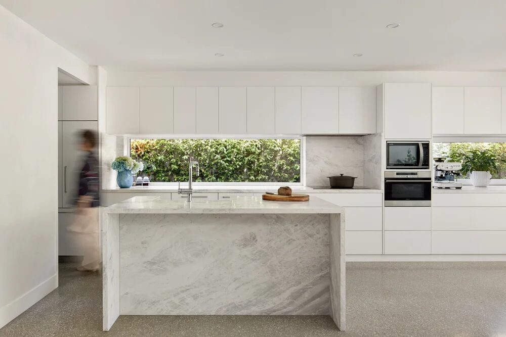 White kitchen with a marble island and a long, low window above the bench looking onto a hedge