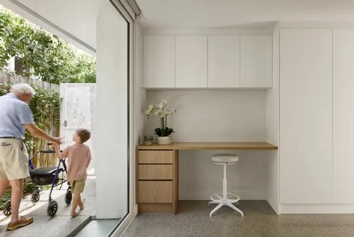 Grandfather with a walker and a young child heading out through a full-height glass door beside a built-in study nook