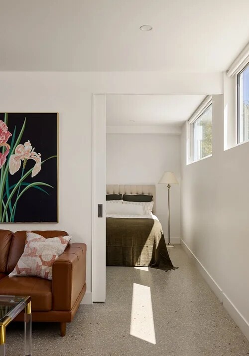 Living room opening through a sliding door to a bedroom lit by high white-framed windows