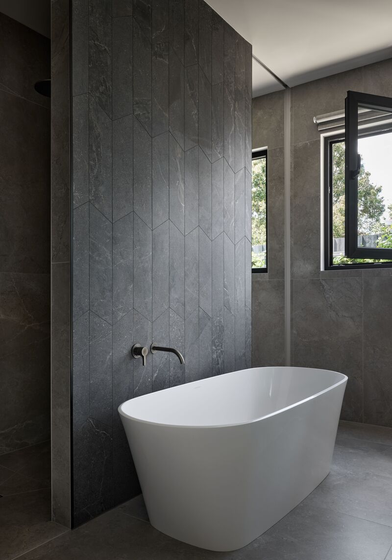 Freestanding bath in a dark tiled bathroom, with a black-framed window open to the garden
