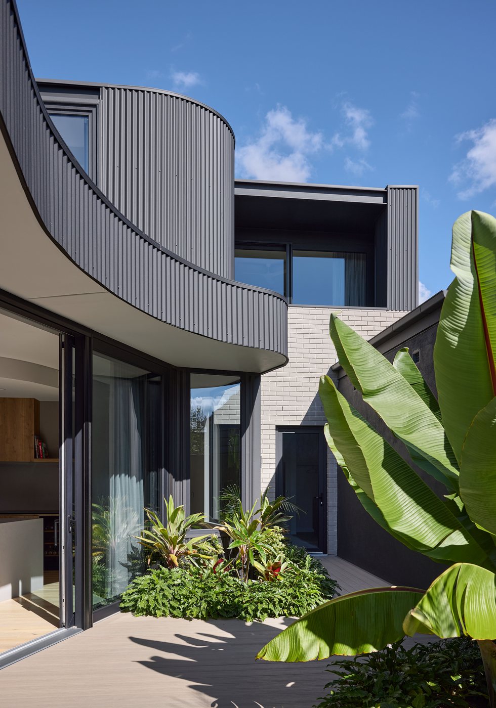 Charcoal uPVC sliding doors and windows beneath a curved corrugated eave, framed by tropical planting